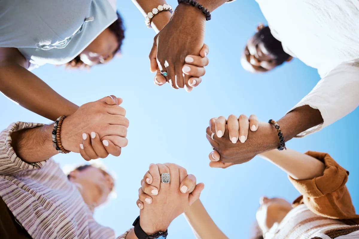Seven people standing in a circle, seen from below against a bright blue sky, each clasping the hands of the two people beside them.